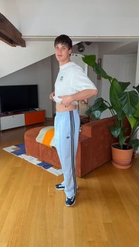 Fiatal férfi fehér pólót emel, hogy megmutassa a hasát, fa padlón áll a nappaliban, ahol narancssárga kanapé, nagy zöld növény és TV-konzol található.