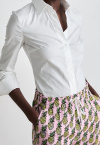 Henkilö, jolla on päällä valkoinen napitettu paita, joka on työntynyt vaaleanpunaisiin housuihin, joissa on keltaisia ja vihreitä ananaskuvioita, kädet taskuissa.