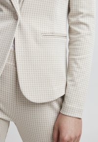 Šviesiai smėlinis švarkas su tekstūruota Houndstooth raštu, užsegamas vienu mygtuku ir su šoniniu kišeniu. Suderintas su atitinkamomis kelnėmis.