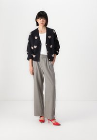 Cardigan nero con motivi a cuore, top bianco, pantaloni a quadro grigi a gamba larga e scarpe con tacco rosso a punta.