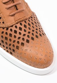 Scarpa in pelle marrone perforata con motivi a diamante e quadrato, suola bianca in gomma, design con lacci e superficie liscia.