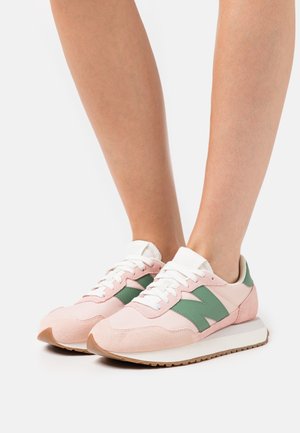 Piedi che indossano sneakers New Balance rosa e verdi con lacci bianchi e suole beige, in piedi su uno sfondo bianco uniforme.