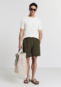 Camicia bianca a maniche corte con texture, pantaloni corti verde oliva e sandali marroni. Porta una borsa a tracolla in tela beige.