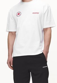 Hvid bomulds t-shirt med korte ærmer, prydet med et cirkulært rødt logo og "PEGADOR" i rødt tekst på forsiden. Udekoreret design.