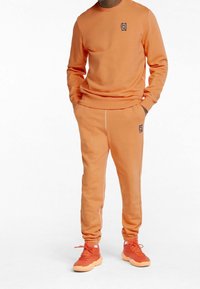 Felpa arancione e pantaloni da jogging abbinati, realizzati in tessuto morbido, con logo ricamato. I pantaloni hanno polsini elastici e cuciture a contrasto.