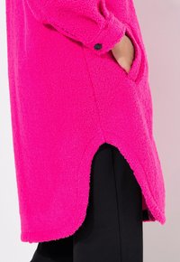 Abrigo de forro polar en un vibrante color rosa con un acabado texturizado. Presenta bolsillos laterales y un dobladillo redondeado. Detalle de puños abotonados visible. Pantalones negros debajo.