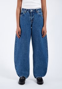Modré oversize džíny z denimu s vysokým pasem, s knoflíkovým zapínáním, širokými nohavicemi a surovým lemem. Nosené s černými botami.