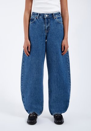 Jean en denim oversize bleu avec taille haute, présentant une fermeture à bouton, des jambes larges et un ourlet brut. Porté avec des chaussures noires.