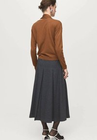 Donna con top a maniche lunghe marrone, gonna midi plissettata grigia scura, collant trasparenti neri, décolleté bianche aperte e orecchini d'argento, di spalle.