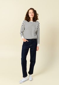 Top de manga larga a rayas en blanco y negro, combinado con jeans de mezclilla azul oscuro. El modelo lleva zapatillas blancas y está de pie contra un fondo beige.