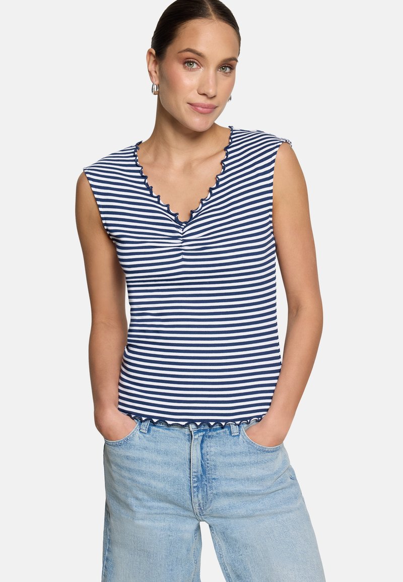 Femme portant un haut sans manches rayé marine et blanc avec un col et un ourlet festonnés, assorti à un jean en denim bleu clair.