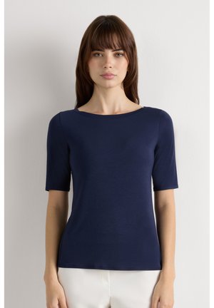 Frau mit glatten braunen Haaren, die ein eng anliegendes marineblaues kurzärmeliges Oberteil und weiße Hosen trägt, steht vor einem einfarbigen hellen Hintergrund.