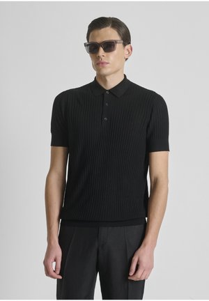 Man met naar achteren gestyled haar, een zwarte geribd kortemouwen polo en zwarte broek tegen een grijze achtergrond, en donkere zonnebril.