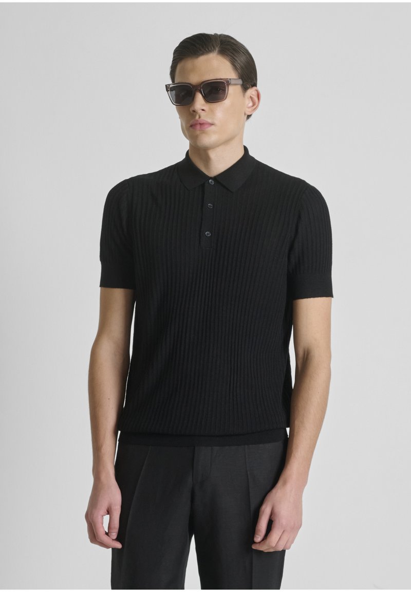 Hombre con el pelo peinado hacia atrás, lleva gafas de sol oscuras, una camiseta polo negra de manga corta con nervaduras y pantalones negros sobre un fondo gris.