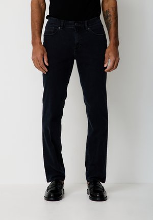 Man wearing black slim-fit jeans, black loafers, and a black top, standing against a plain white background.