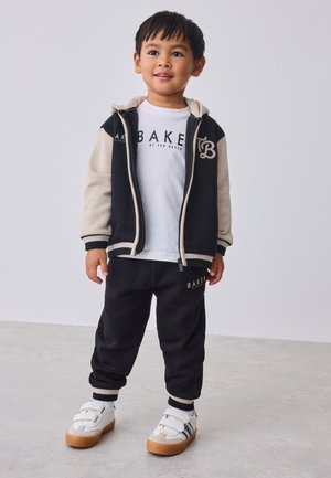 Ragazzo giovane in piedi che indossa una giacca con cappuccio nera e beige, una maglietta bianca con la scritta "BAKER", pantaloni neri e sneaker bianche.