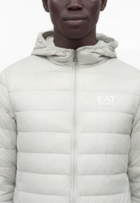 Homme portant une veste matelassée gris clair avec une fermeture éclair frontale et une capuche, arborant le logo blanc EA7 Emporio Armani sur la poitrine.