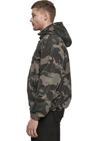 Sötétzöld és szürke árnyalatú kamuflázs dzseki, kapucnival, rugalmas mandzsettákkal, cipzáras oldalsó zsebekkel és laza fazonnal.