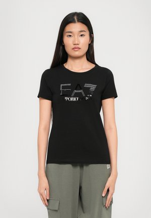 Zwarte t-shirt met korte mouwen, voorzien van een glanzend logo met rhinestones; ronde halslijn en zacht, glad materiaal. Gecombineerd met olijfgroene broek.