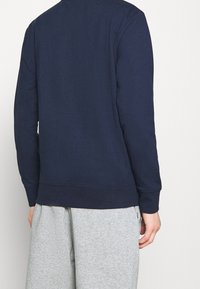 Sweat-shirt bleu marine à col rond et ourlet côtelé, doté de manches longues et d'une texture lisse. Porté avec un pantalon de survêtement gris.