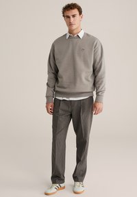 Sivá mikina s okrúhlym výstrihom a logom, vrstvená cez bielu košeľu s golierom. Hnedé kárované nohavice a svetlomodré tenisky dopĺňajú outfit.