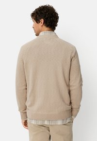 Beige sweater with a ribbed hem and cuffs, rounded neck, and subtle texture. Worn over a gray and white plaid shirt.