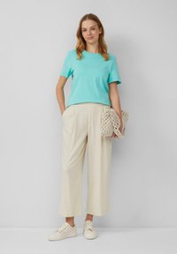T-shirt in cotone turchese chiaro, vestibilità rilassata, abbinato a pantaloni larghi color crema. Il modello tiene in mano una borsa in rete beige e indossa sneakers bianche.