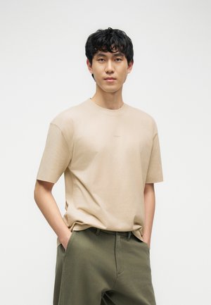 Noor mees, lühikeste mustade juustega, kandes beeži t-särki ja oliivrohelisi pükse, seisab käed taskutes kogu aeg ühevärvilise tausta ees.