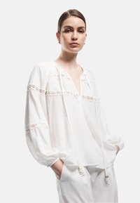 Blusa bianca con maniche lunghe, dettagli in pizzo, scollo a V e chiusura con cordoncino. Tessuto morbido e strutturato con una vestibilità comoda e accenti sottili.