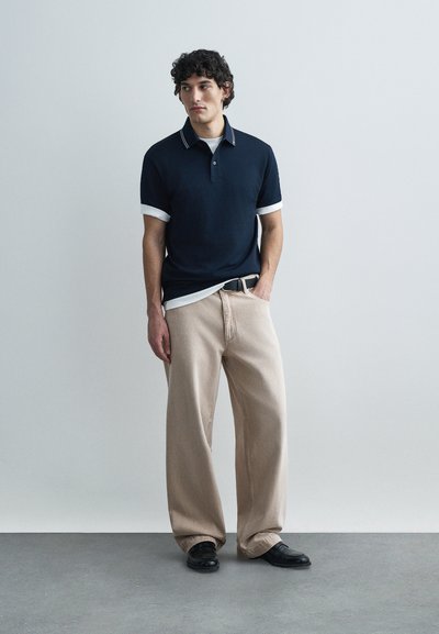 Μπλε ναυτικό πόλο πουκάμισο με λευκά μανσέτα, συνδυασμένο με μπεζ παντελόνι φαρδιάς γραμμής και μαύρα παπούτσια. Το μοντέλο στέκεται μπροστά σε ένα ανοιχτό γκρι φόντο.