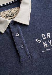 Marineblauw poloshirt met witte kraag, drie donkere knopen, geborduurd met "S.D.R N.Y" en een binnenlabel met de tekst "Superdry Co."