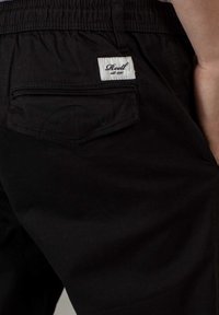 Pantalones cortos de algodón negros con una cinturilla elástica, un bolsillo trasero con un detalle de costura curva y una etiqueta blanca cosida en la tela.