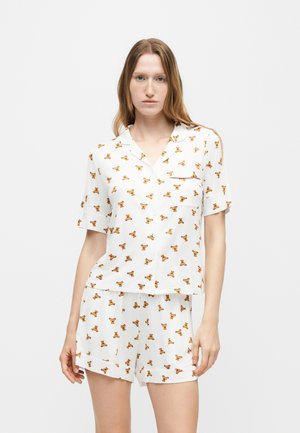 Femme portant un haut blanc à manches courtes boutonné et un short assorti avec un motif de petits ours en peluche orange.