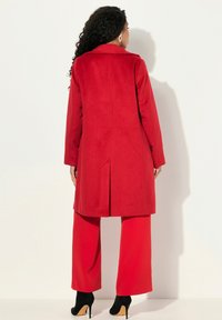 Rode wollen jas met een op maat gemaakt ontwerp, lange mouwen en een rugsplit. Gecombineerd met rode broek met wijde pijpen en zwarte stiletto's.