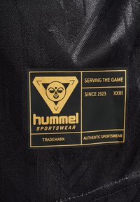 Svart tygetikett med guldtext och en triangeldesign med en logotyp. Det står "hummel SPORTSWEAR," "SERVING THE GAME," och "SINCE 1923."