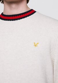 Pull crème avec un col côtelé aux rayures rouges et navy. Logo jaune brodé sur la poitrine. Tissu en tricot texturé.