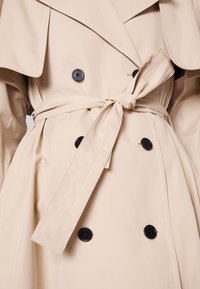 Trench-coat beige avec des boutons noirs et une ceinture nouée à la taille, montrant la partie médiane et une partie des manches.