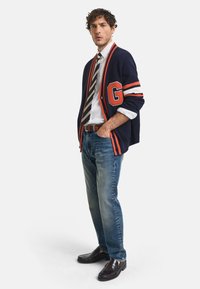 Marineblauer Cardigan mit rot-weißen Streifen und einem "G"-Patch, getragen über einem weißen Hemd, gestreifter Krawatte, blauen Jeans und dunklen Slippern.