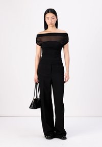 Čierny top bez ramienok z priesvitnej látky, skombinovaný s čiernymi nohavicami s vysokým pásom. Modelka drží malú čiernu kabelku s minimalistickým dizajnom.
