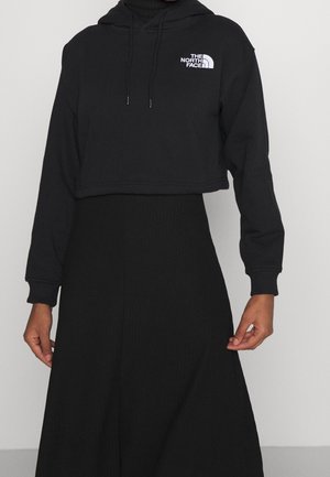 Krótka czarna bluza z kapturem z logo "The North Face" na piersi noszona na długą czarną spódnicę, ręce zrelaksowane wzdłuż boków.