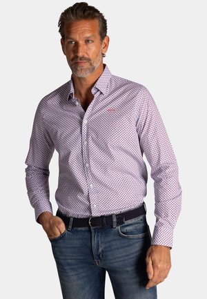 Man met grijze baard, gekleed in een patroon lange mouwen shirt dat in een blauwe spijkerbroek met zwarte riem is gestopt, staand met één hand in zijn broekzak.