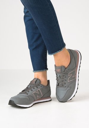 Personne portant des baskets New Balance grises avec des touches roses et un jean bleu foncé à ourlet effiloché, debout sur un sol blanc.