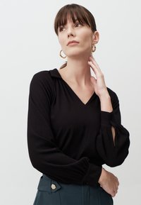 Haut noir à manches longues côtelé, avec un décolleté en V et des poignets boutonnés en forme de trou de serrure. Le tissu est doux, avec un léger éclat.