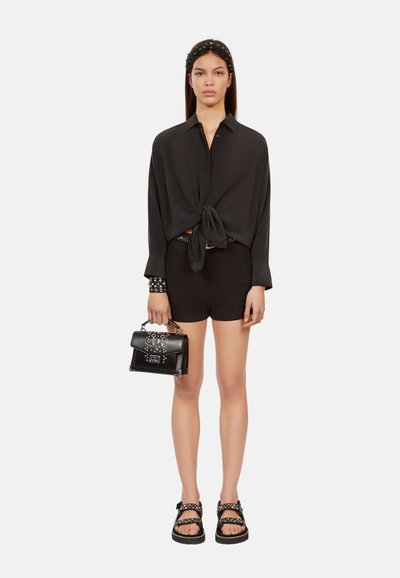 Zwarte blouse met strik, lange mouwen, hoge taille zwarte shorts en zwarte sandalen met studs. Houdt een kleine zwarte handtas met zilveren accenten vast.