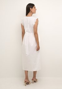 Žena stojící bokem v bílé midi šatech bez rukávů s vyšívanými detaily a béžovými vysokými podpatky.