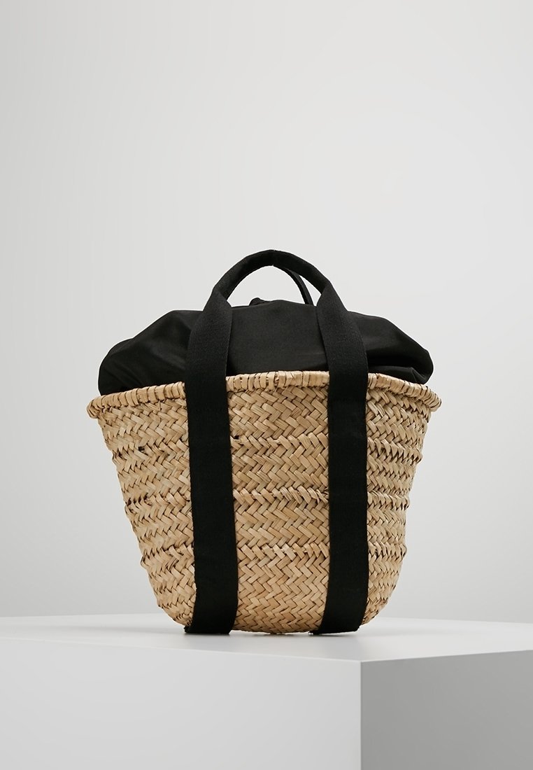 Borsa a tracolla in paglia intrecciata con manici in tessuto nero e chiusura superiore in tessuto nero, esposta su un piedistallo bianco contro uno sfondo grigio.