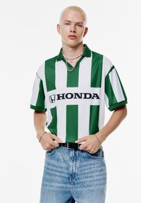 Groen en wit gestreepte polo met kraag, voorzien van een zwart "HONDA" logo. Draag het met lichtblauwe, gestructureerde denim jeans.