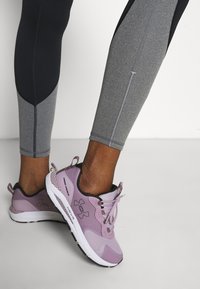 Lila träningsskor med nät- och syntetöverdrag, med en vit sula och svarta accenter. Bärs med gråa och svarta leggings.