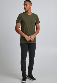 Krátké rukávy tričko olivově zelené barvy s horizontálními pruhy, s náprsní kapsou, spojené s černými úzkými džíny a tmavými teniskami.