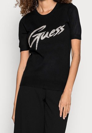 Woman wearing a black short-sleeve top with the word "Guess" in large rhinestones across the chest.